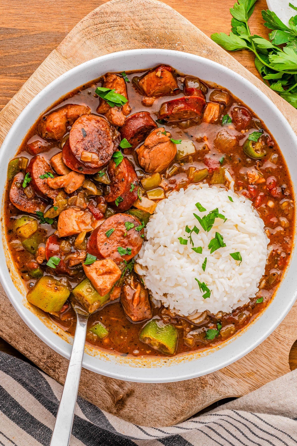 A bowl of gumbo with sausage, chicken, vegetables, and a serving of white rice, garnished with chopped parsley. A bowl of gumbo with sausage, chicken, vegetables, and a serving of white rice, garnished with chopped parsley.