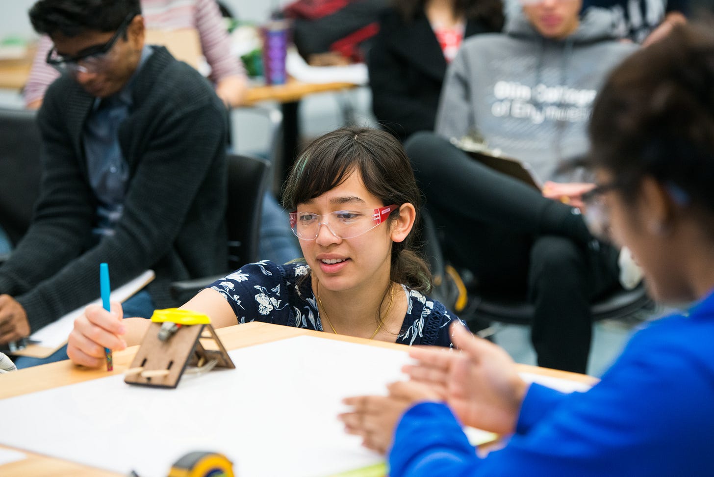 In an engineering laboratory, a student and professor confer about a small mechanical prototype on a table. The student wears safety glasses and is kneeling to get a closer view. In the background, other students look on.