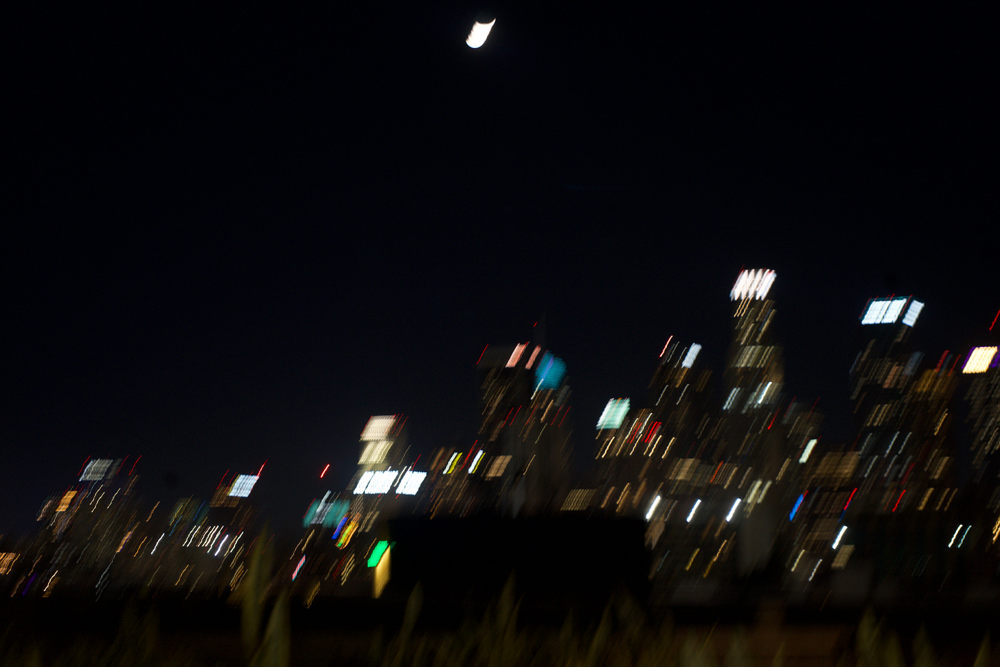 Softly blurred nighttime city skyline with glowing building lights and a crescent moon in the dark sky above. Softly blurred nighttime city skyline with glowing building lights and a crescent moon in the dark sky above.