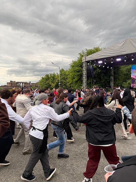 Photos from the Concours Mondial de Bruxelles cultural program in Armenia, including a group dinner at a long candlelit table, a meeting room with the Armenian minister of economy beneath the national flag, a display of Soviet-era wine industry memorabilia and archival photos, aging wine barrels in a cellar, a group photo overlooking Yerevan with Mount Ararat in the distance, illuminated performers at the Garni Temple at night, traditional dancers performing acrobatics, a crowd joining a folk dance circle outdoors, and a large karas amphora covered in visitor signatures." Let me know if you'd like it trimmed or reordered to lead with a particular image.