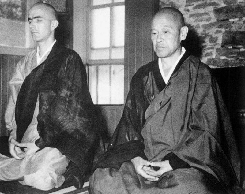 Foto em preto e branco de Richard Baker (à esquerda) e Suzuki-roshi (à direita) em mantos tradicionais budistas, sentados em meditação.