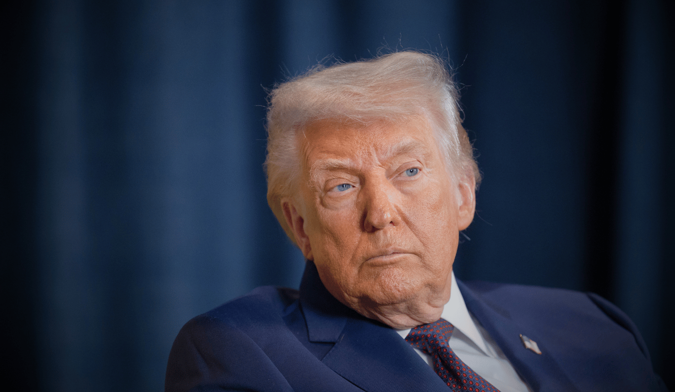 Donald Trump in navy suit and red tie, seated against blue curtain backdrop, looking to one side.
