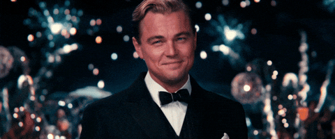 Scena dal film Il grande Gatsby. Un uomo elegante in smoking (interpretato da Leonardo DiCaprio) sorride leggermente mentre alza un calice di champagne per brindare. Sullo sfondo, luci scintillanti e fuochi d'artificio creano un'atmosfera festosa.