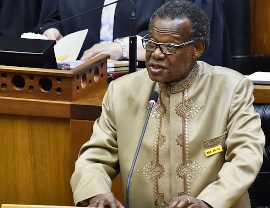 An elderly, bespectacled man speaks at a podium 