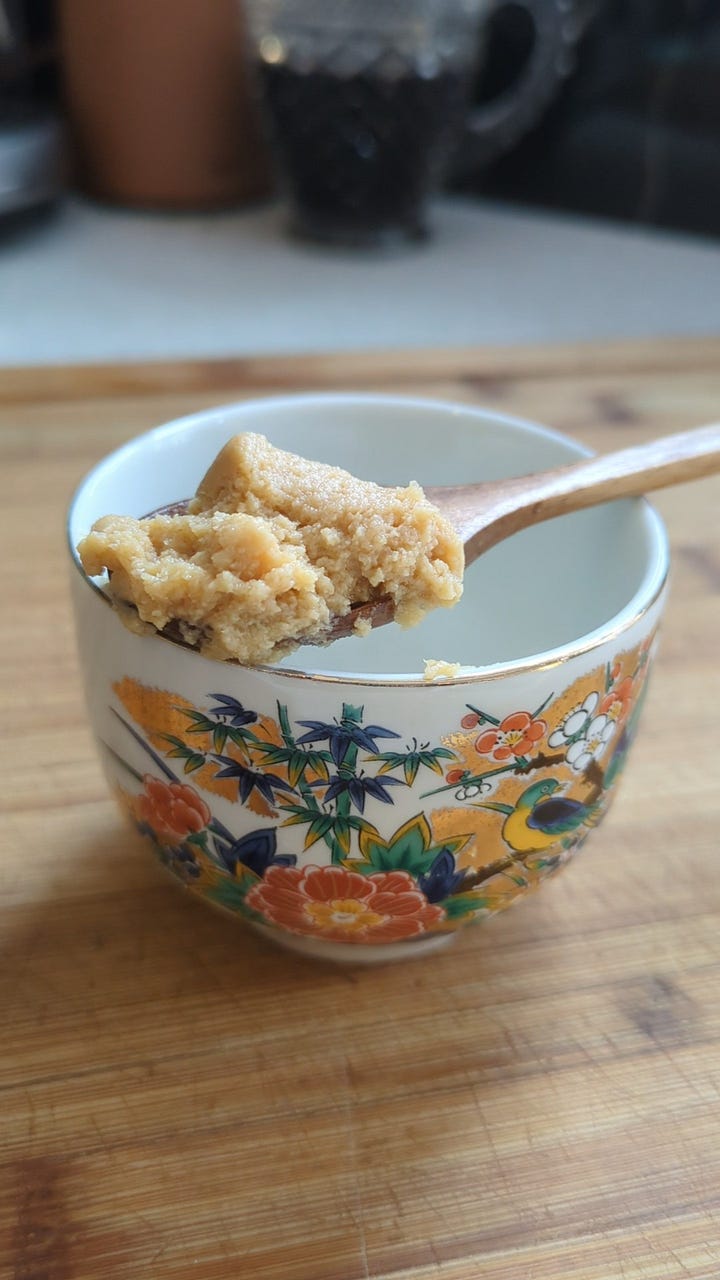 delicate teacup with asian art, with a small wooden spoonful of miso tare. Golden spoonful of fresh home made chicken and duck ramen broth spilling into glass container.