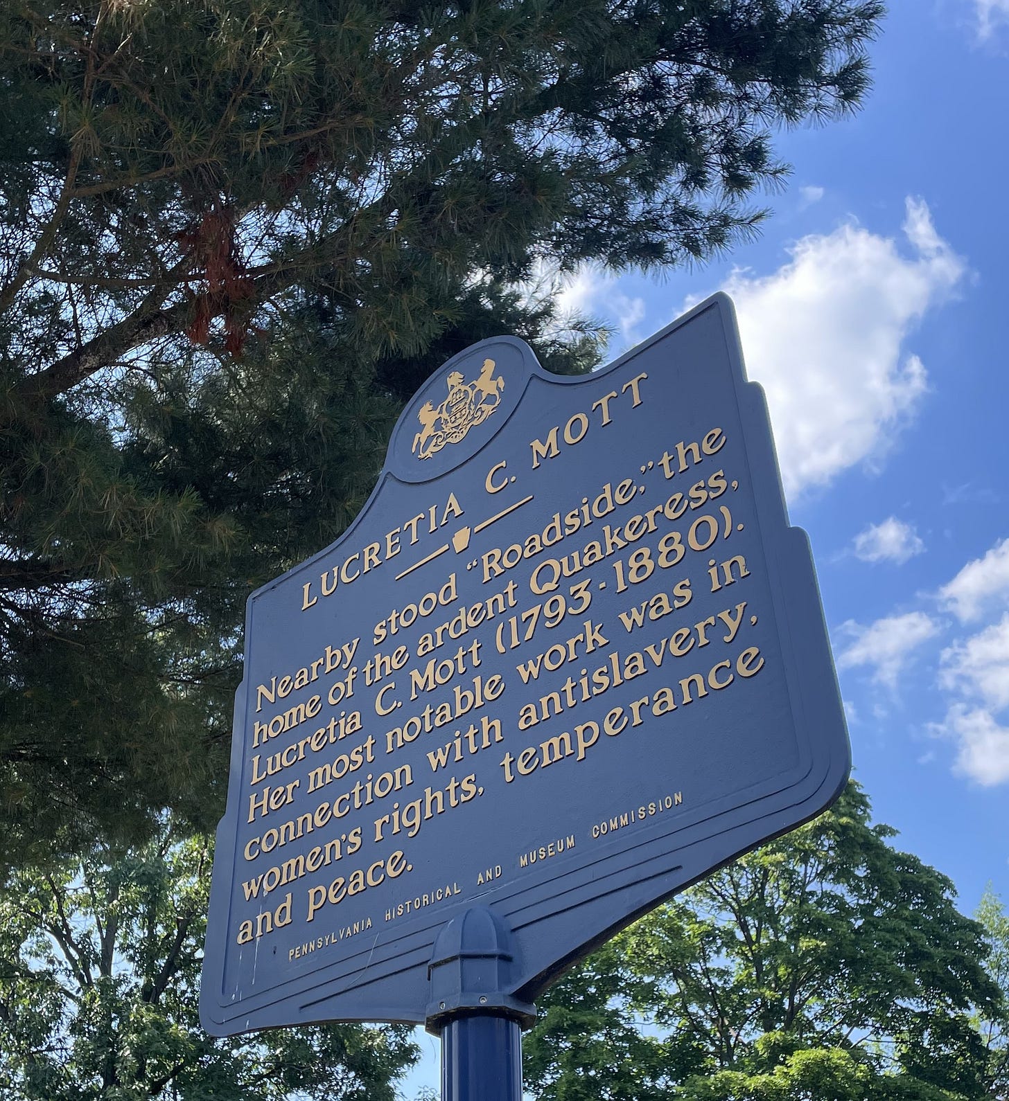 Blue historical marker indicating nearby location of Lucretia Mott's home, Roadside, with blue sky and leafy tree branches in background. Blue historical marker indicating nearby location of Lucretia Mott's home, Roadside, with blue sky and leafy tree branches in background.