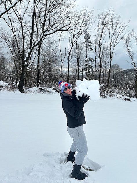 Woman in a Bills beanie and black jacket holds a small snowball, and then a basketball-sized snowball, and then stands next to a giant snowball that is almost as tall as her.