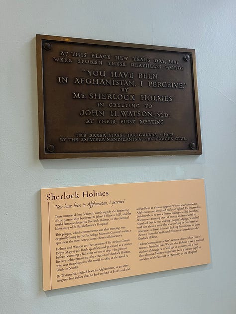 The door to the Barts Museum, a plaque commemorating Holmes and Watson's first meeting, and a large wooden sign advertising medical care.