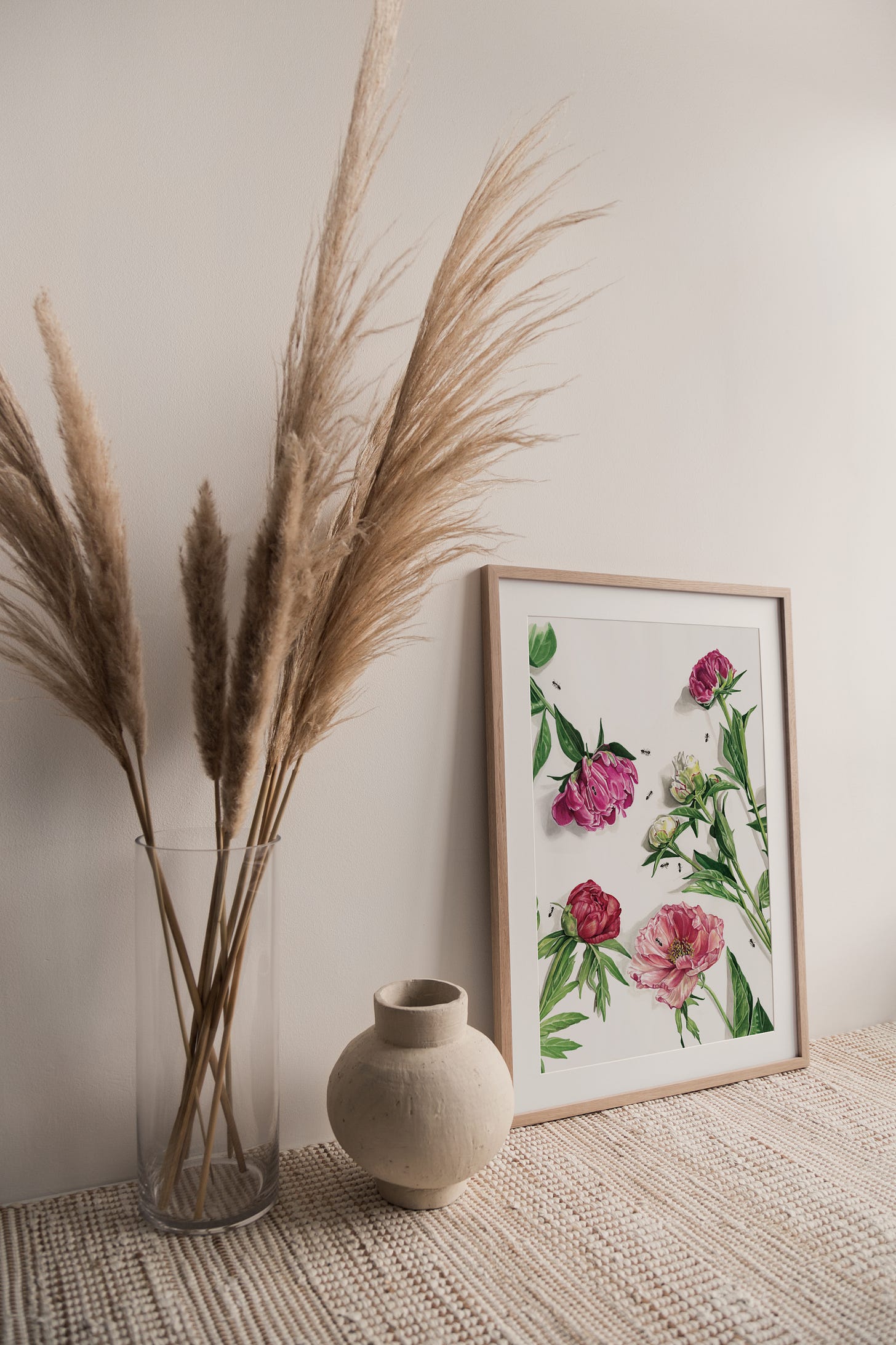 An image of a vase with dried flowers and a framed print of the peony painting to the right of the vase, propped up against the wall at an angle. 