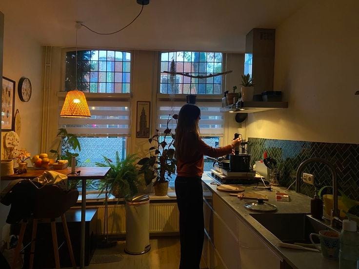 This may contain: a woman standing in a kitchen next to a sink and window with plants on the windowsill