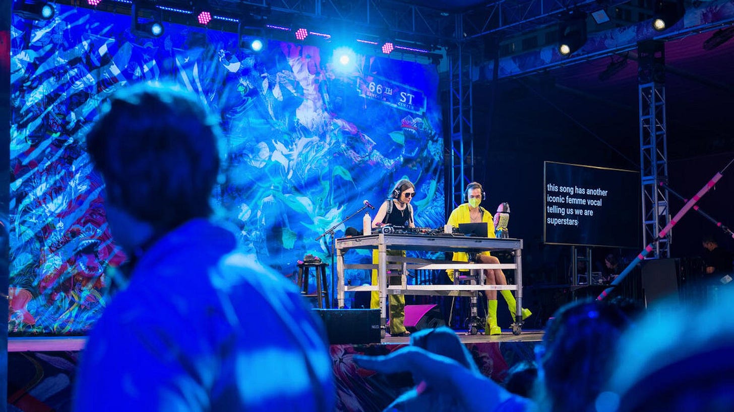 a photo from a darkened dance floor toward a stage with a dj at work against a collage of images from summer in new york city. another person is working at a computer, typing captions on a screen nearby: this song has another iconic femme vocal telling us we are superstars.