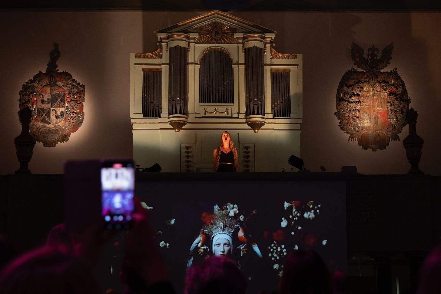 A dark phtotgraphic image from the inside of a grand building. A young white woman is underlit dramatically, dwarfed by the interior. Her mouth is open in song or scream. Below her a video image of a beaufiul myserious woman is illuminated. Someone has raised their phone to take a photo and the screen is blurry in the foreground