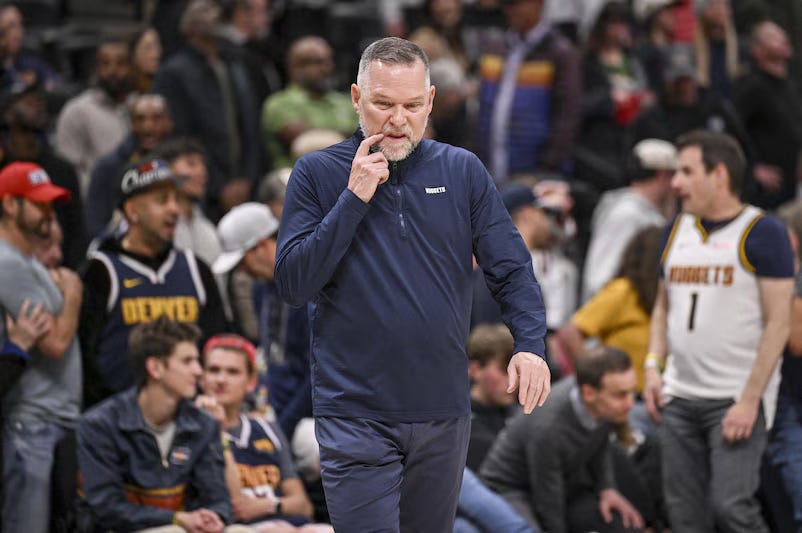 Basketball coach in navy jacket deep in thought on sideline during Denver Nuggets game with crowd and bench players in background