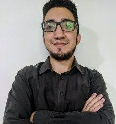 Foto de Alyson Gomes olhando para a câmera, com um sorriso e braços cruzados. Alyson tem barba, usa óculos e uma camisa de botão cinza escuro. Foto de Alyson Gomes olhando para a câmera, com um sorriso e braços cruzados. Alyson tem barba, usa óculos e uma camisa de botão cinza escuro.