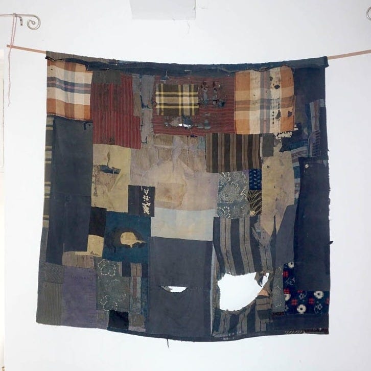May include: A large, patchwork textile hanging on a white wall. The textile is composed of various fabric squares in shades of blue, brown, and beige. Some squares have patterns like stripes and plaid. The textile has visible wear and tear, with some holes.