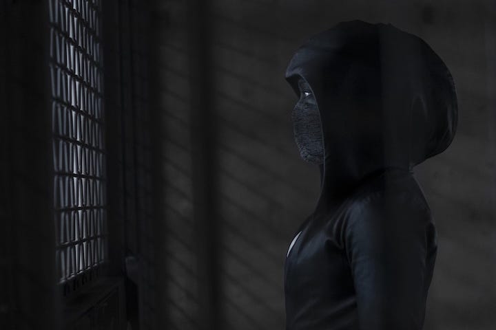 Regina King as Sister Night, clad in a hooded leather outfit and face mask, stands in profile behind a grated window, bathed in low, moody light.