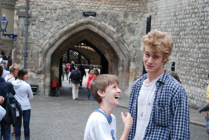 Standing outside the Tower of London, one tall teen boy makes a silly face while a shorter boy laughs uncontrollably.