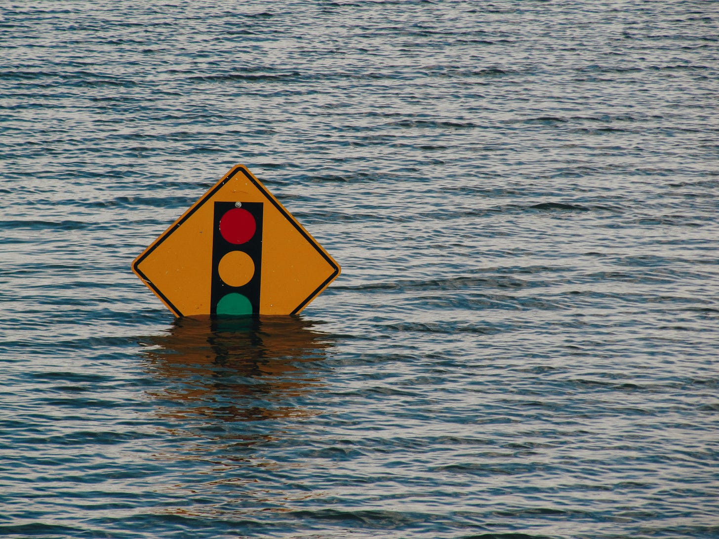 Warning sign depicting a traffic light partially submerged in floodwater, with the water reaching halfway up the sign, indicating severe flooding.