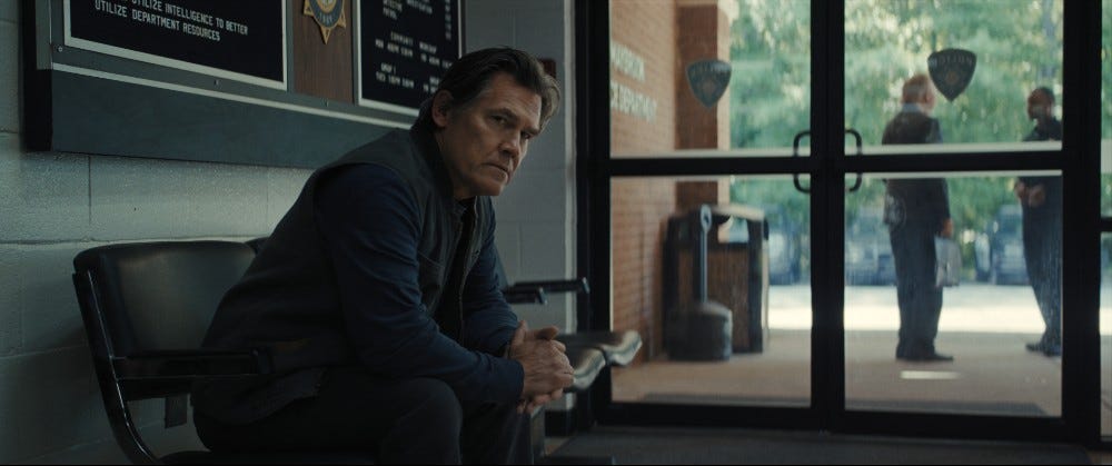Josh Brolin in Weapons, sitting in a police station with two officers standing outside the glass door.