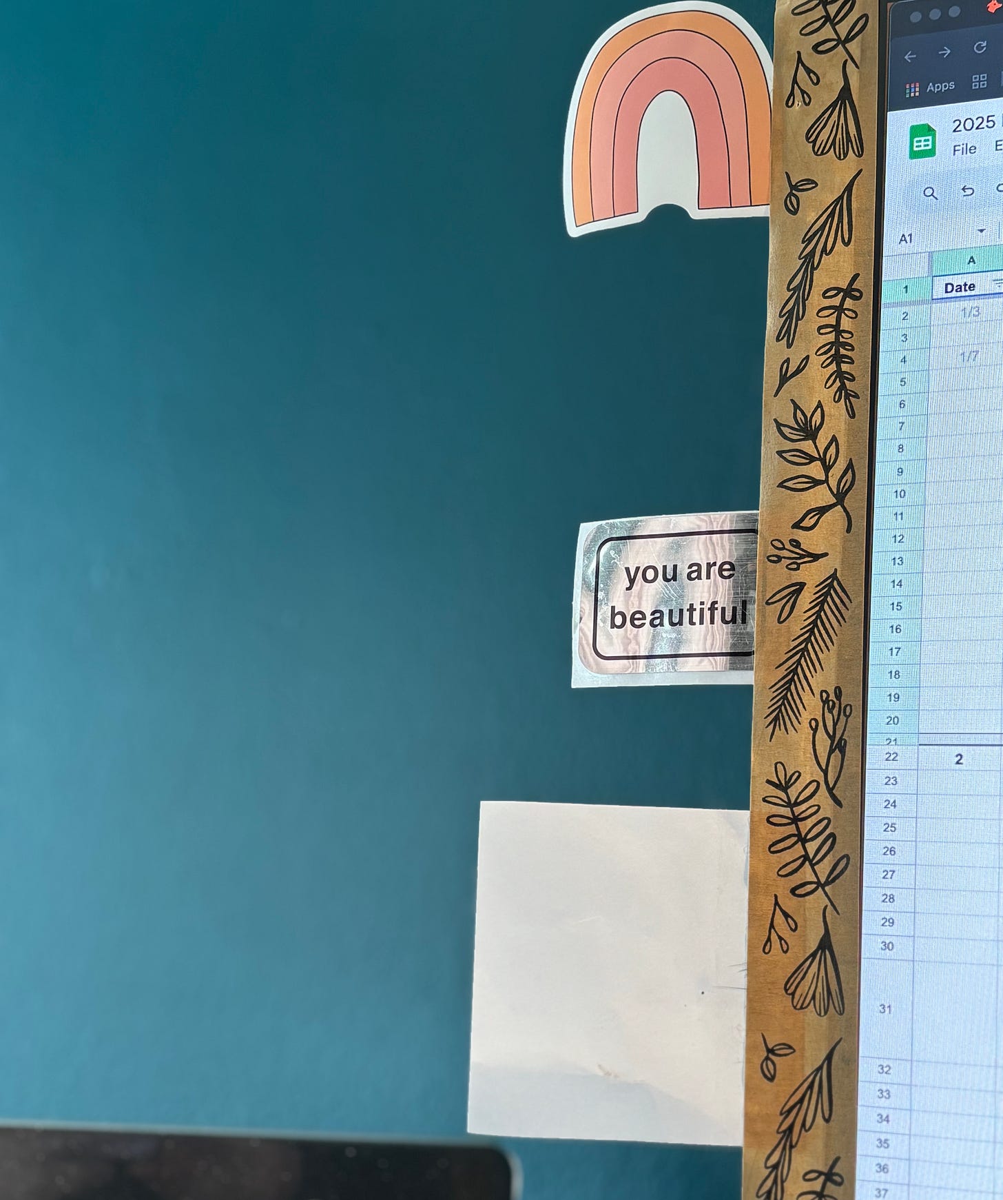 Floral tape on the side of a monitor holds (from the top down) a stylized rainbow sticker, a shiny silver "you are beautiful" sticker, and a blank white post-it note.