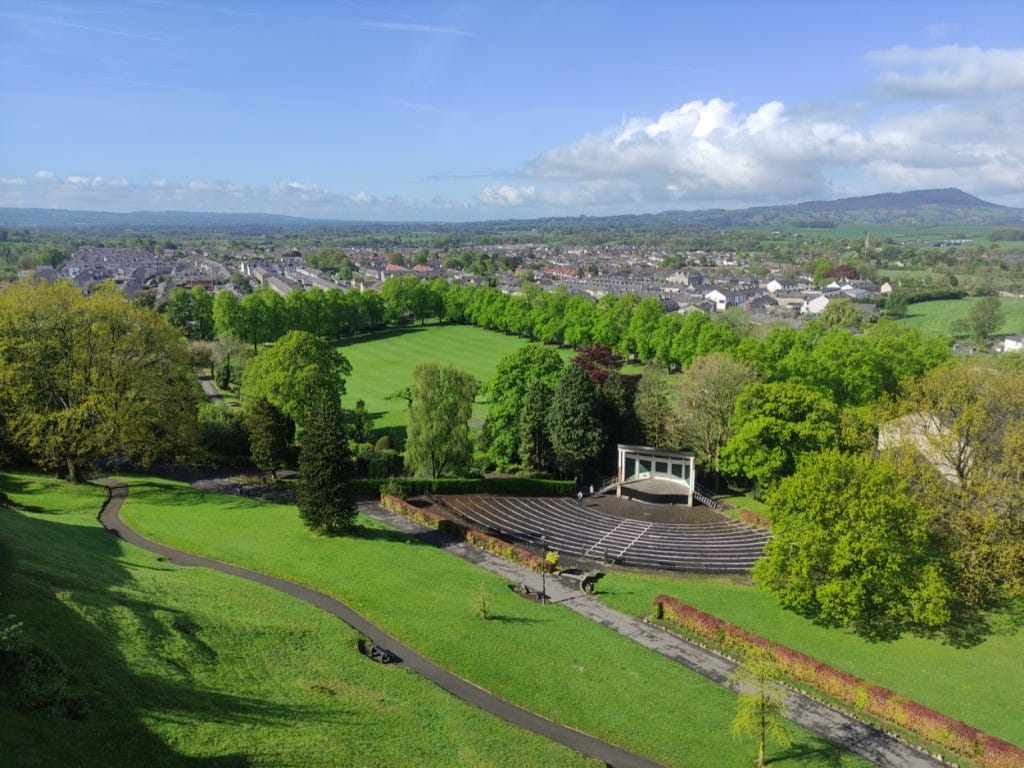 Looking down from the height of the castle, seeing a path winding below, the whole of the bandstand and its concrete steps, and houses behind the well-defined tree-lined border of the park.