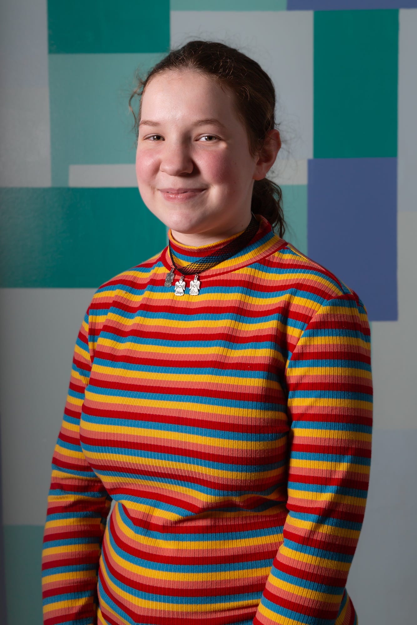 A 13 year old white skinned girl wearing a multicolored striped turtle neck. She has reddish brown hair and light brown eyes. Her hair is pulled back into a ponytail. She is smiling at the camera.