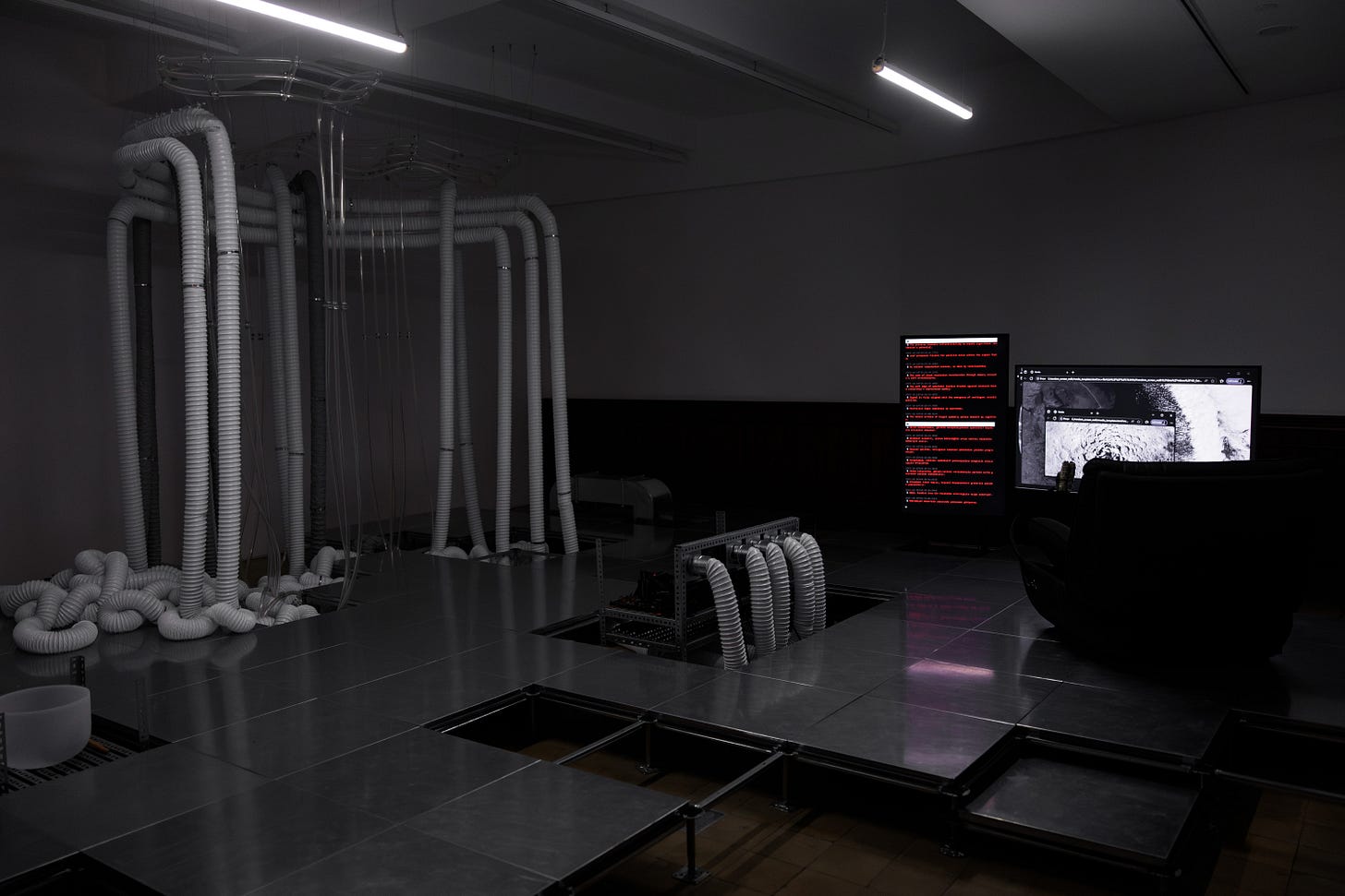 A dark gallery space dimly lit by some fluorescent light tubes. On show are white corrugated tubing running from the ceiling to the floor, resembling some sort of tentacled being. On a raised metal platform are some large digital displays are computer programming code with red text on black background and a second computer display showing a planetary landscape. A dark gallery space dimly lit by some fluorescent light tubes. On show are white corrugated tubing running from the ceiling to the floor, resembling some sort of tentacled being. On a raised metal platform are some large digital displays are computer programming code with red text on black background and a second computer display showing a planetary landscape.