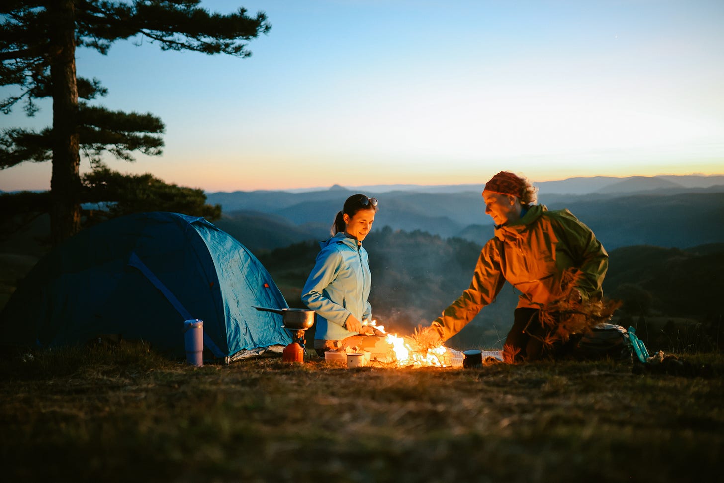Two people cooking over a fire at dusk next to a tent Two people cooking over a fire at dusk next to a tent