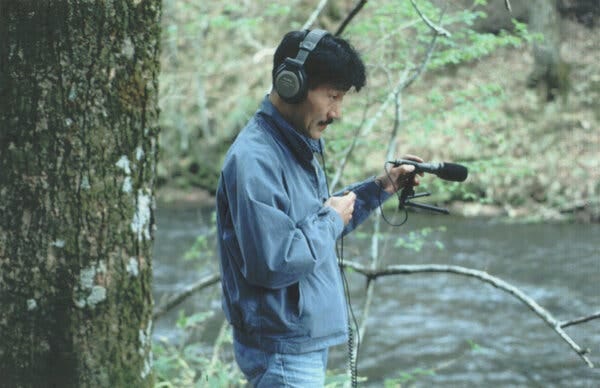 A man wearing headphones and a windbreaker stands next to a stream, holding up a microphone toward the water.