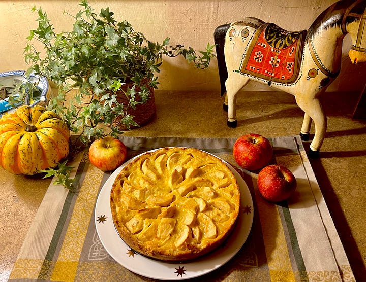 A four-photo collage: an apple tree heavy with small red apples in tall grass; a hand reaching up to pick a red apple from a branch; a golden apple tart on a white plate beside loose apples, a small striped squash, ivy and a painted wooden horse; and two bowls and a cloth tote bag full of freshly picked apples and pears on a terracotta floor.