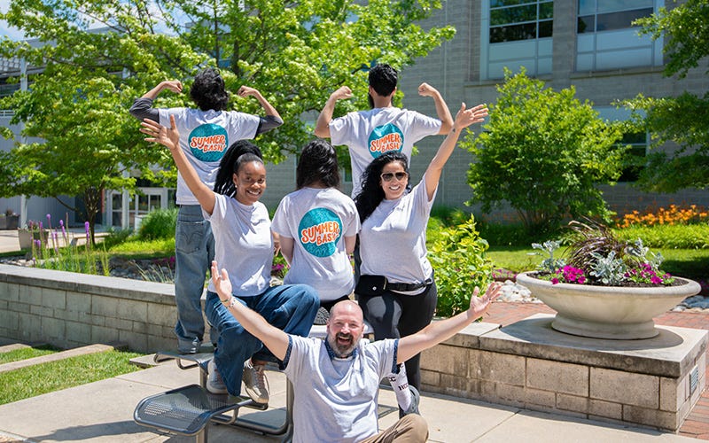 "Six Howard Community College students and staff in matching 'Summer Bash' T-shirts pose playfully outdoors on a sunny campus courtyard. Several face away to show the logo on the back of their shirts, arms raised in celebration, with green trees, flowering plants, and a brick planter in the background."