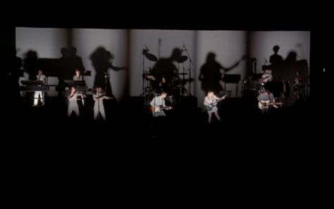 selects from Talking Heads' concert film Stop Making Sense. images of Byrne holding a microphone to camera, raising his arms, dancing in a big suit an with a lamp and full shots of the band.