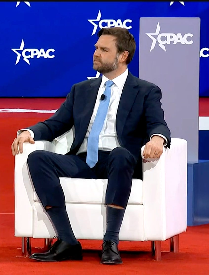 JD Vance sits in a white chair on the CPAC stage. He is wearing a light blue tie that waterfalls down over his crotch, and skinny-leg suit pants. Sitting down has made the pantlegs hike up o JD Vance sits in a white chair on the CPAC stage. He is wearing a light blue tie that waterfalls down over his crotch, and skinny-leg suit pants. Sitting down has made the pantlegs hike up o