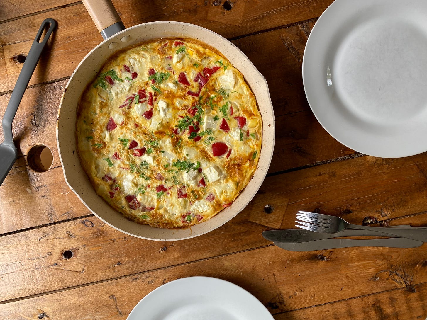 Frying pan with omlette. Red flecks and white cubes of cheese are showing in the omelette. The pan with plates and cutlery is on a wooden table. 