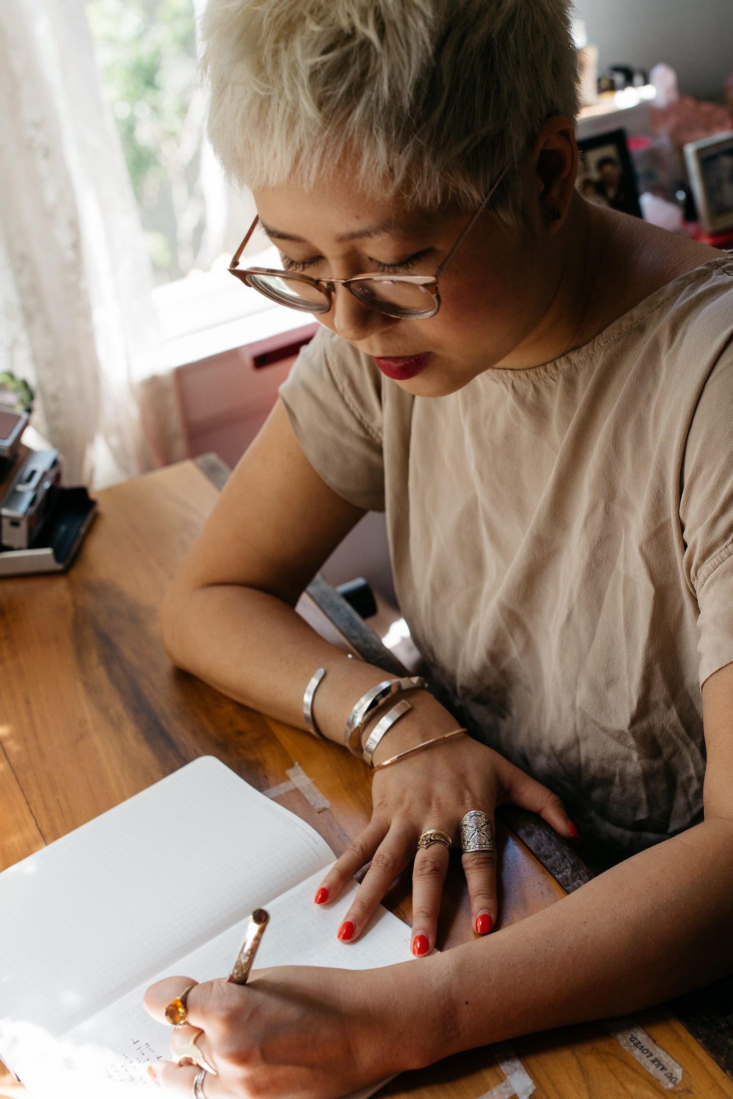 A woman with short platinum blonde hair and glasses writes in a notebook at a wooden desk. She wears multiple rings, bangles, and red nail polish. Sunlight filters through a window beside her.