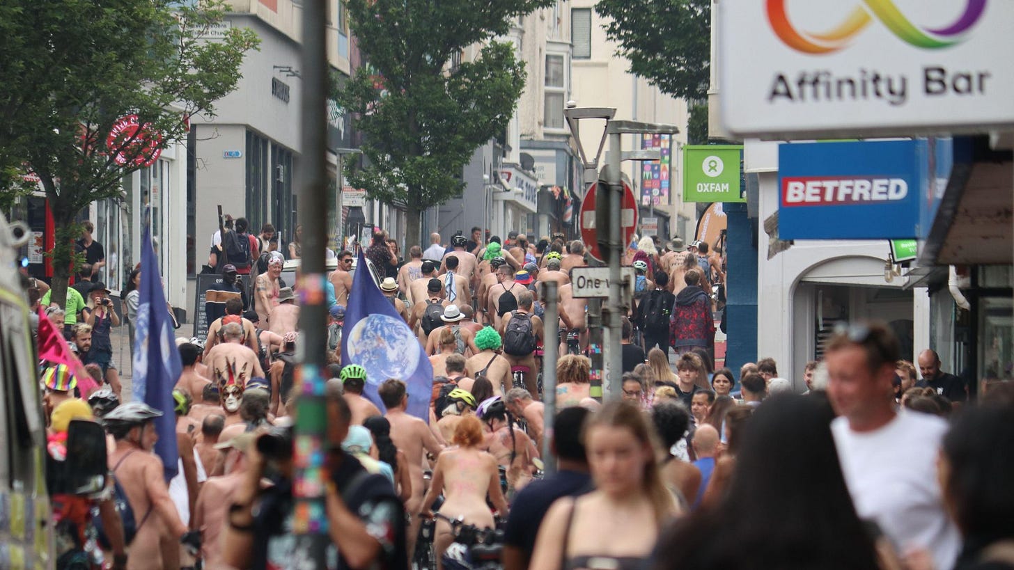 Hundreds of nude cyclists ride through a crowded street in Brighton during the World Naked Bike Ride, surrounded by pedestrians, storefronts, and colorful flags. Hundreds of nude cyclists ride through a crowded street in Brighton during the World Naked Bike Ride, surrounded by pedestrians, storefronts, and colorful flags.