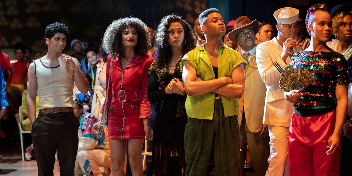 In a crowded room of people, the viewer is focused on a forward-facing line of seven BIPOC queer people wearing stylish colorful clothing and looking to the right as they all watch a ballroom performance happening offscreen. 