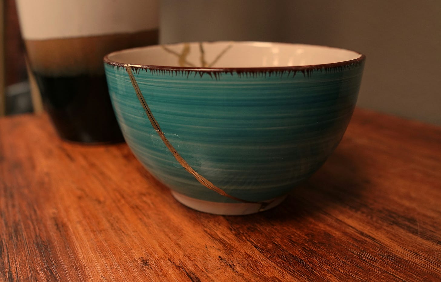Close-up of a turquoise ceramic bowl repaired with golden cracks in the Japanese Kintsugi style, sitting on a warm wooden table with another ceramic vessel blurred in the background. Close-up of a turquoise ceramic bowl repaired with golden cracks in the Japanese Kintsugi style, sitting on a warm wooden table with another ceramic vessel blurred in the background.
