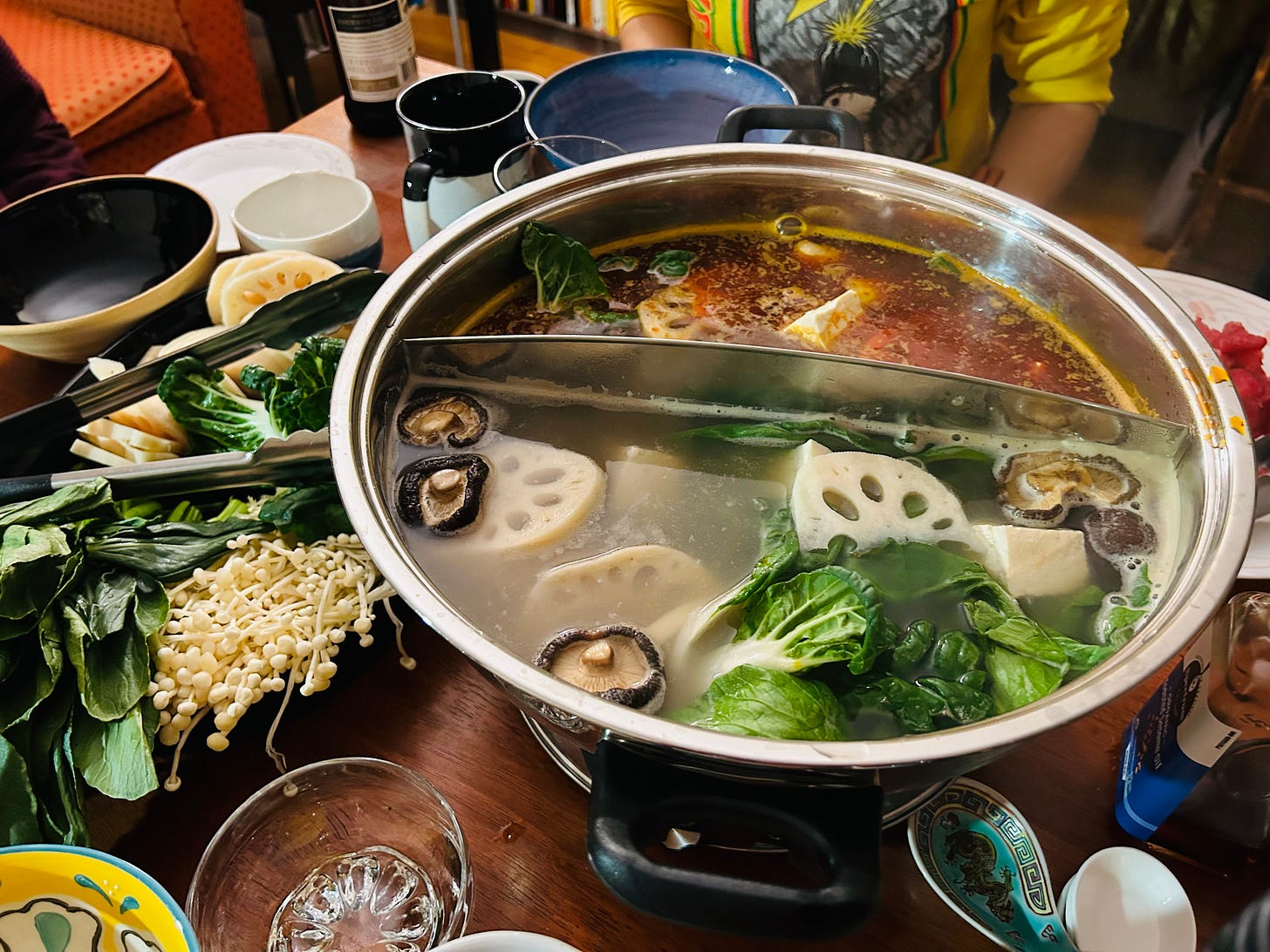 A big metal pot holding two kinds of broth is surrounded by plates of vegetables and bowls. A big metal pot holding two kinds of broth is surrounded by plates of vegetables and bowls.
