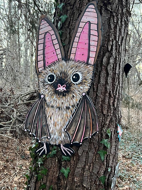 First image is a picture of an upright painted bat with pink ears hanging on a tree, second image is the entry way to the Batland Exhibit + Save the Woods party, next is a painted sign on fabric that reads KEEP ASHEVILLE GREEN, fourth is an image of the Batland Exhibit sign with Spencer Beals, my daughter, and me, fifth is a close up of the Batland Exhibit sign which is a map of the woods showing where the bats are placed- sixth is an image of a party goer with large homemade see- through bat wings posing in front of the sign, sevent is a home made painted sign that reads 'Chancellor to Helene "Hold My Beer! Watch Me Take All the Trees!" eight is an image of Spencer's painting titled "Eyes of Appalachia" and finally we have a hand made carrier pigion on a tree as part of the exhibit.