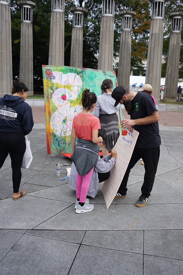 A woman holding a white bag examines a brightly colored painted giant book. A child uses a marker to add to a book made out of pillows and furniture. A group of people examines a colorful giant book with painted text and images. A giant, brightly colored book stands in front of a building with columns.
