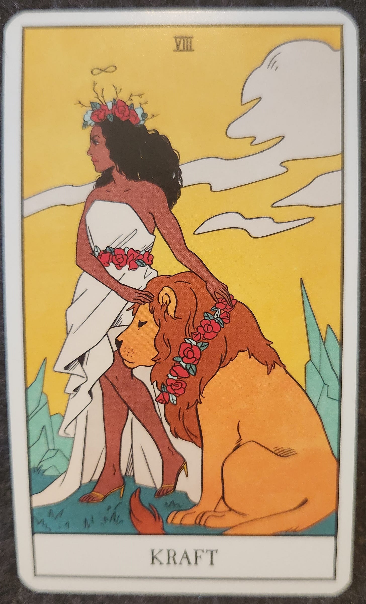 A tarot card labelled XIII Kraft. A woman is facing the left of the card, wearing a long white tunic dress with a belt of flowers around her waist. Next to her a large lion is sitting. She is petting the lion with both hands. Both the woman and the lion are wearing a crown of roses. In the background is a line of trees and a yellow sky with white clouds. 