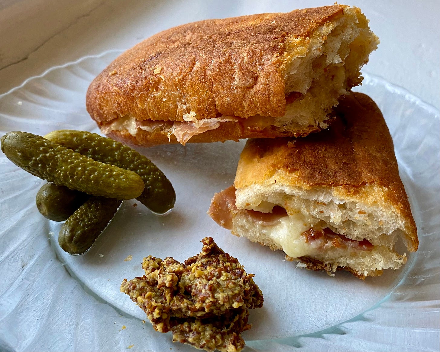 grilled cheese on a baguette with prosciutto, with cornichons and seedy mustard on the side, presented on a clear glass plate on a white countertop