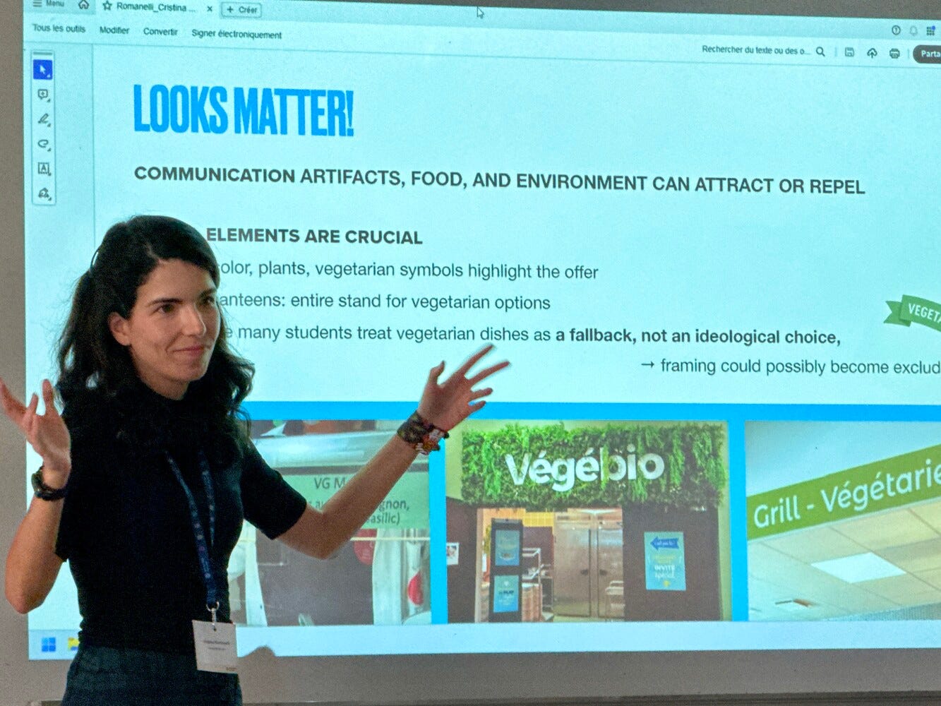 Woman in front of screen with slide. Text: LOOKS MATTER! Communication artifacts, food, and environment can attract or repel. Some photos on the screen show different versions of vegetarian labeling in student canteens in France. 