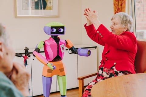 A small bright multicoloured humanoid robot interacts with an elderly woman wearing a red jacket A small bright multicoloured humanoid robot interacts with an elderly woman wearing a red jacket