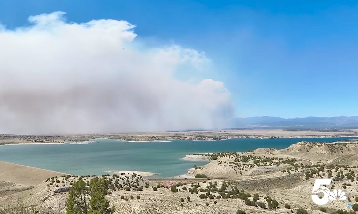 Smoke crosses a blue sky over a water ringed desert lake.