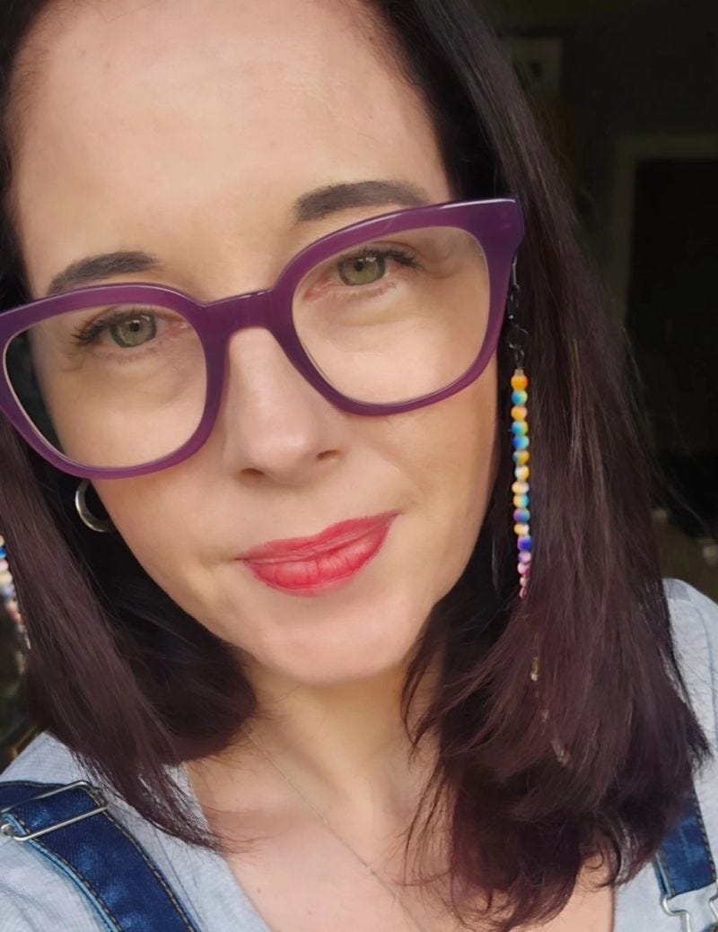 Portrait of a woman with shoulder-length dark hair wearing purple glasses and a grey top under denim overalls. She is looking directly at the camera with a gentle, natural expression. A small heart-shaped necklace is visible, and the photo is taken in soft natural light indoors.