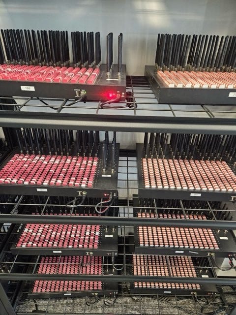 Multiple black SIM servers with numerous SIM card slots, some with red indicators, arranged on metal racks in a room. Several SIM servers and tangled cables scattered on a wooden floor in a room with a window, dark curtains, and a door. No watermarks from platforms like Instagram, TikTok, or Xiaohongshu are present.