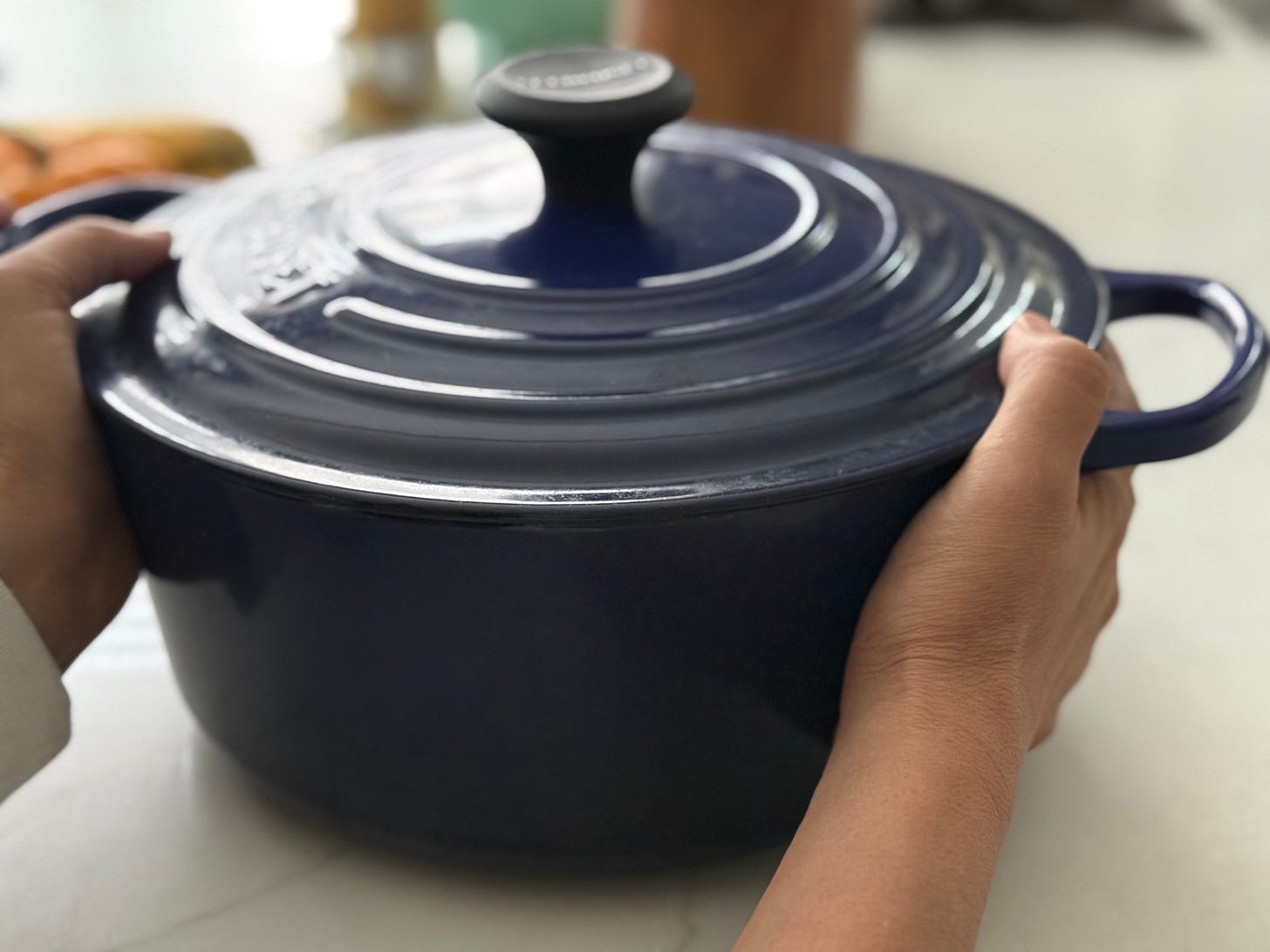 Hands cradling a navy enameled Dutch oven with the lid on, resting on a kitchen counter.