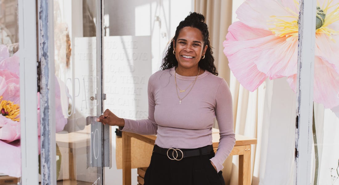 Tarnya Bruce - a woman wearing a pink shirt and black jeans - opening the door to her studio, with pink flowers painted on glass panels either side of the walls